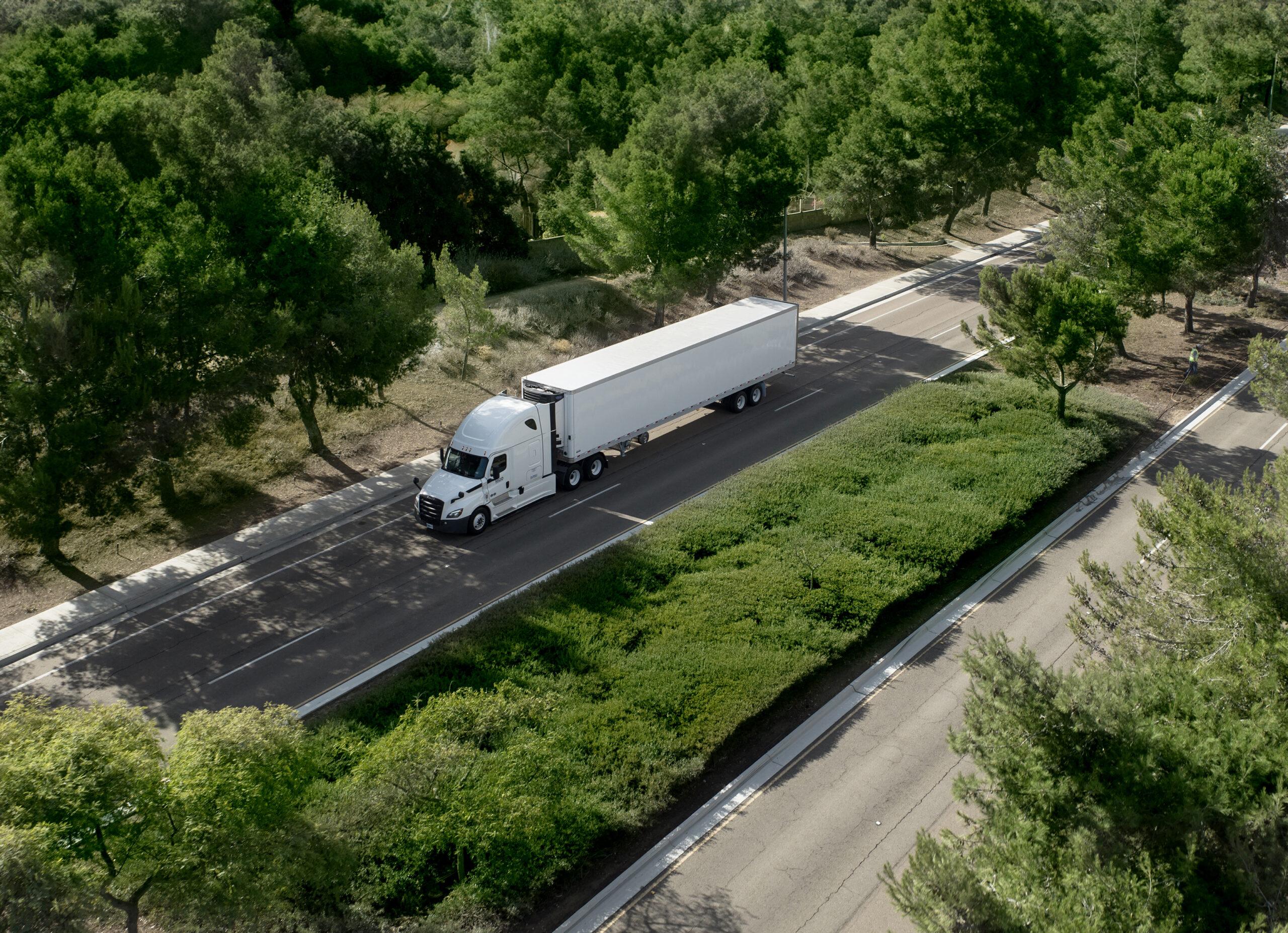 DIN6868_1337 An aerial view of a white Freightliner Cascadia semi-truck pulling a Hyundai Translead trailer on a road surrounded by trees, demonstrating Hyundai's trailer innovation and thermal efficiency technology.