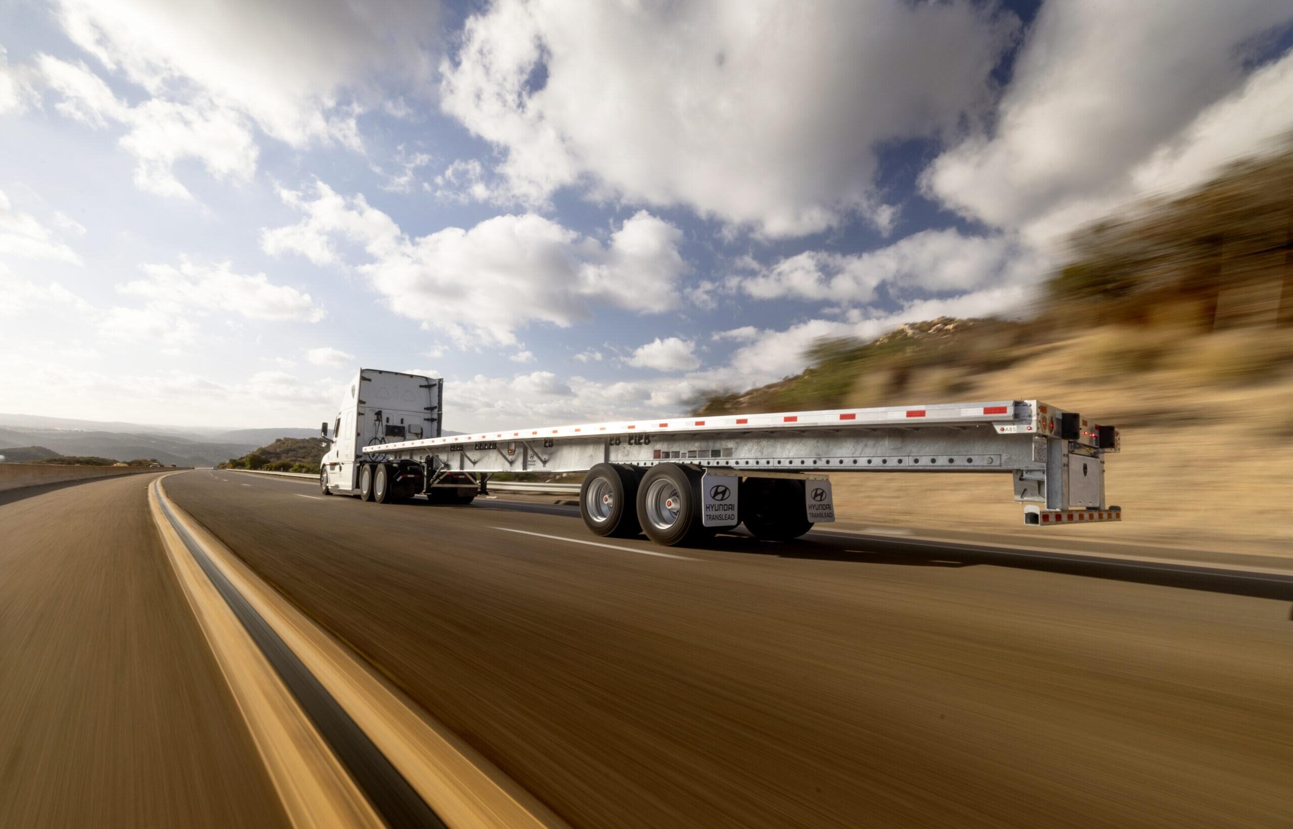 A Hyundai Translead Latitude Flatbed trailer being pulled by a semi-truck on a highway with a hilly landscape, showcasing Hyundai's innovation and flatbed trailer technology in motion.