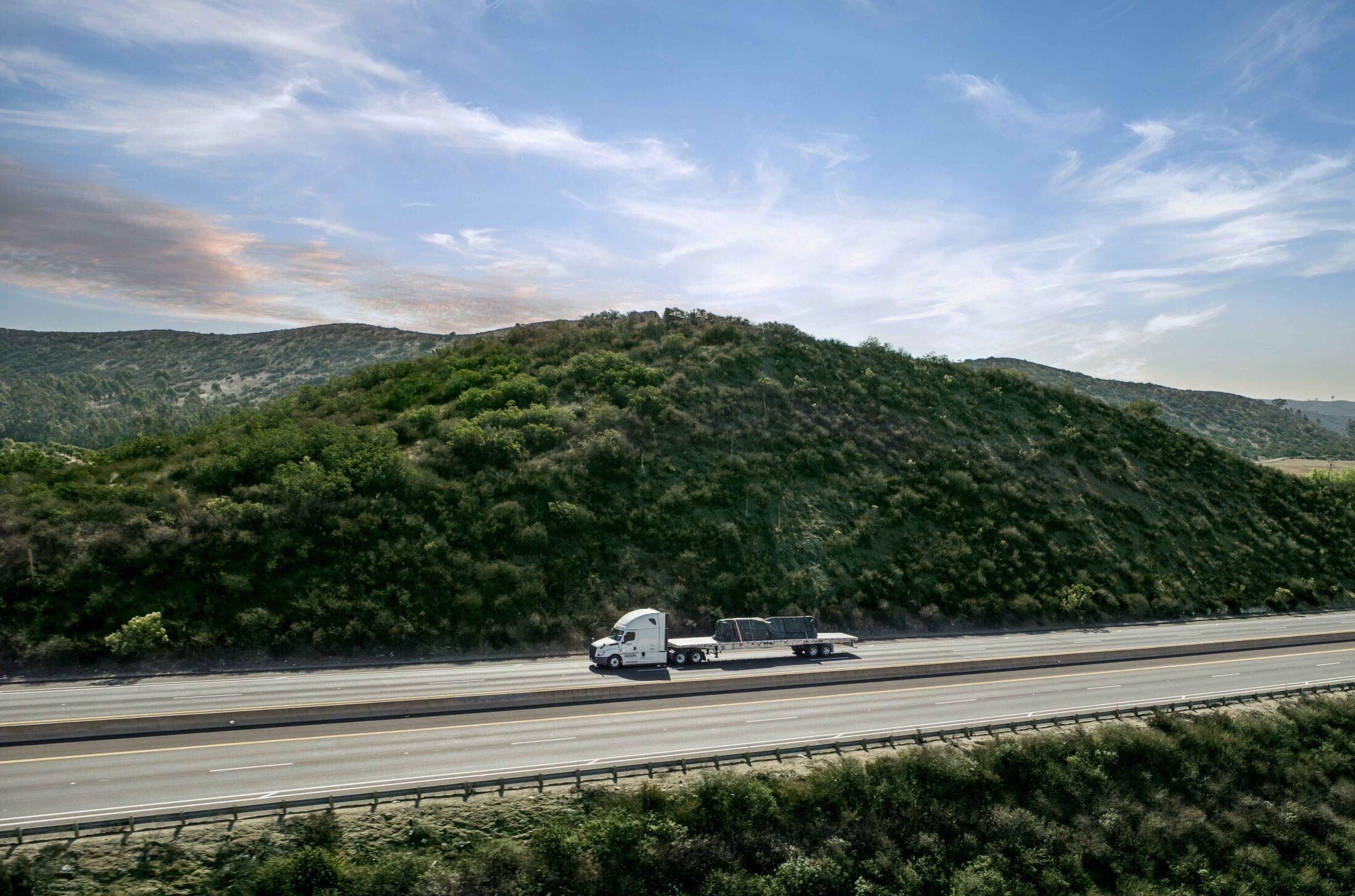 An aerial view of a semi-truck pulling a Hyundai Translead Latitude Flatbed trailer on a highway through green hills, showcasing Hyundai's trailer technology in diverse landscapes.