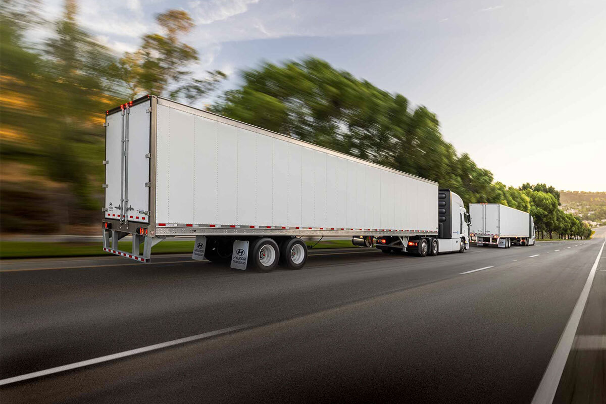 mobile-history Two white Hyundai Translead dry van trailers being pulled by semi-trucks on a highway with blurred green trees, showcasing Hyundai's advanced trailer technology and innovation in transportation.